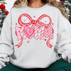 May include: A light gray sweatshirt with a large red bow and the word "MOM" in a heart-themed design. The letters are decorated with pink hearts and patterns. Small pink hearts are scattered around the design.
