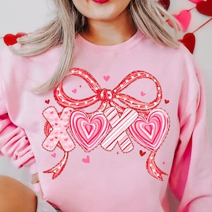 May include: Pale pink sweatshirt with a red bow and the letters "XOX" in a heart design. The design includes hearts and a ribbon, ideal for Valentine's Day. The sweatshirt is a light pink colour.