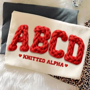 May include: A cream-colored pillow with the letters "ABCD" in bold, red, knitted yarn. Below the letters, the words "KNITTED ALPHA" are embroidered in red, with a heart symbol on each side. The pillow is on a dark gray surface.