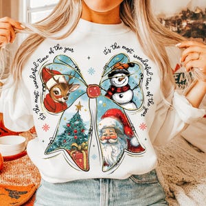 May include: White sweatshirt featuring a large bow design with Christmas-themed illustrations. The bow includes a reindeer, snowman, Christmas tree, and Santa Claus. Text around the bow reads "It's the most wonderful time of the year."