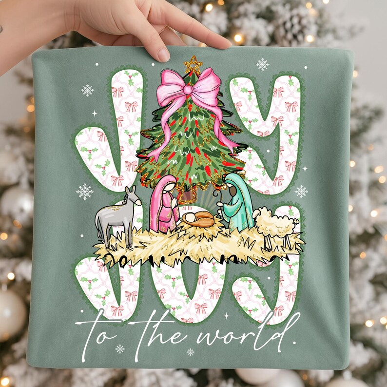May include: A sage green pillow with the words "JOY" and "To the world" in white script. The pillow features a Christmas tree with a pink bow, a nativity scene, and snowflakes. The design is festive and seasonal.