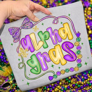 May include: A light gray t-shirt featuring the words "Alpha Gras" in colorful, bubble letters. The design includes a purple bow, gold fleur-de-lis, and a string of beads in green, purple, and gold. The overall theme is Mardi Gras.