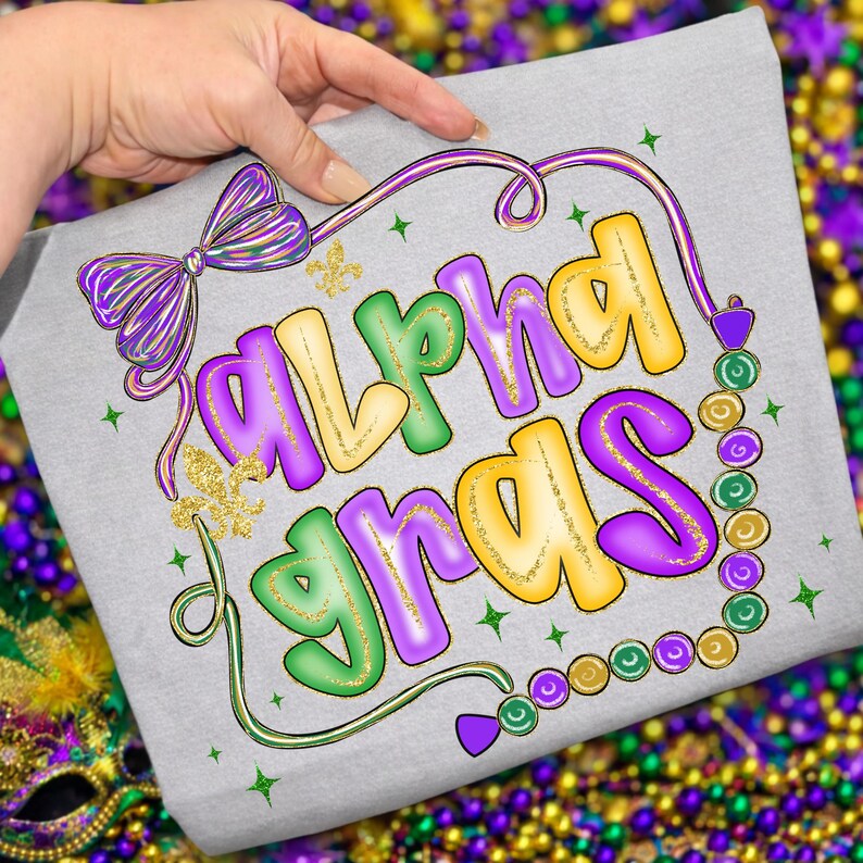 May include: A light gray t-shirt featuring the words "Alpha Gras" in colorful, bubble letters. The design includes a purple bow, gold fleur-de-lis, and a string of beads in green, purple, and gold. The overall theme is Mardi Gras.