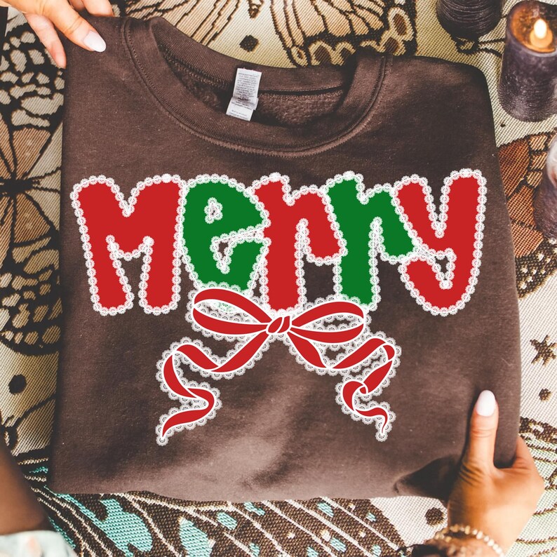 May include: A brown sweatshirt with the word "Merry" in red and green, outlined in white lace. A red bow with white lace detailing is below the word. The sweatshirt is held by hands.