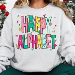 K&ouml;nnte beinhalten: Ein hellgraues Sweatshirt mit den Worten "HAPPY ALPHABET" in bunten Blockbuchstaben. Die Buchstaben sind schwarz umrandet und mit Rosa, Gelb, Gr&uuml;n und Blau gef&uuml;llt. Das Design umfasst Herzen, Sterne und florale Akzente.