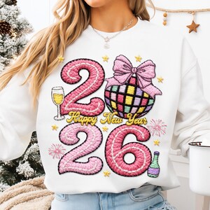 May include: White sweatshirt with a festive design for the New Year. Features the numbers "26" in pink, a disco ball with a pink bow, a champagne glass, and the words "Happy New Year".