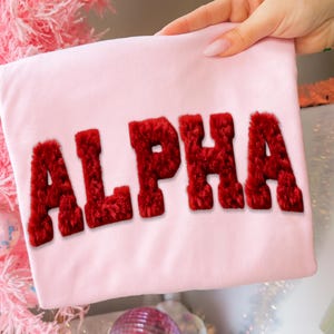 May include: A light pink t-shirt with the word "ALPHA" in bold, red, fuzzy letters. The shirt is held up by a hand with manicured nails. The background includes a pink Christmas tree and decorative items.
