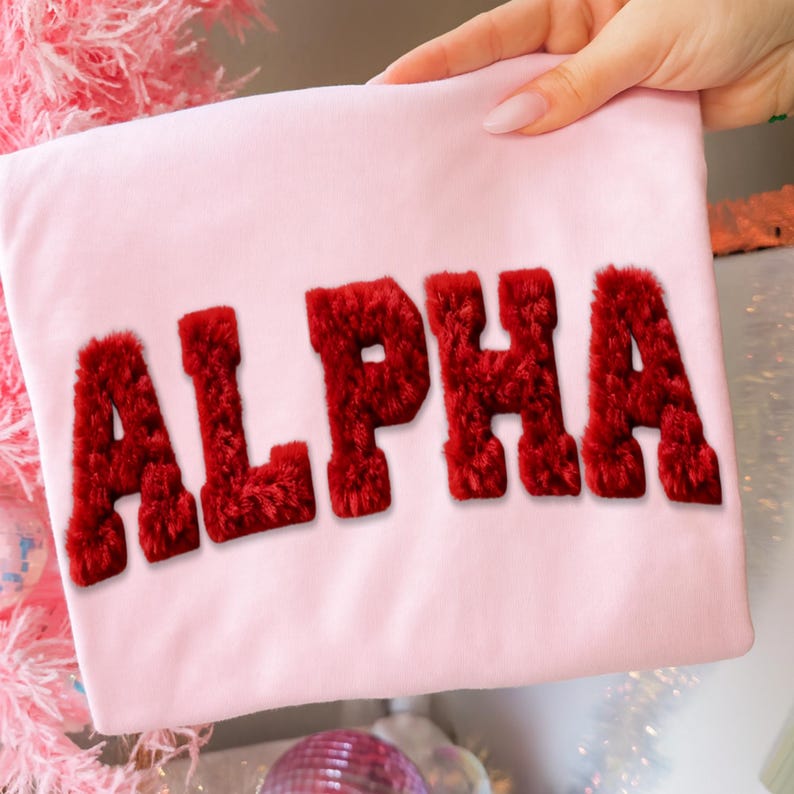 May include: A light pink t-shirt with the word "ALPHA" in bold, red, fuzzy letters. The shirt is held up by a hand with manicured nails. The background includes a pink Christmas tree and decorative items.