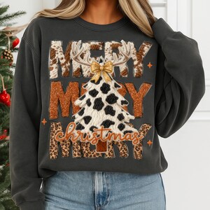 May include: Dark gray sweatshirt with a festive Christmas design. The design features the words "MERRY CHRISTMAS" with a cow print Christmas tree, antlers, and a gold bow. The sweatshirt is paired with blue jeans.