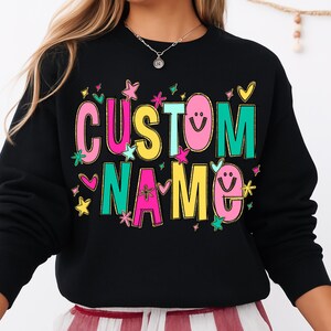 May include: Black sweatshirt with the words "CUSTOM NAME" in colorful, playful lettering. The text is accented with stars, hearts, and smiley faces. The design has a gold outline.