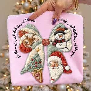 May include: Pink sweatshirt with a large bow design featuring embroidered Christmas elements: a reindeer, snowman, Christmas tree, gift, and Santa Claus. The text "It's the most wonderful time of the year" is arched around the bow.