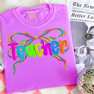Op de afbeelding: Een lila t-shirt met het woord "Teacher" in kleurrijke bubbelletters. Een regenboogstrikontwerp staat boven het woord. Het shirt is opgevouwen en wordt op een oppervlak getoond.