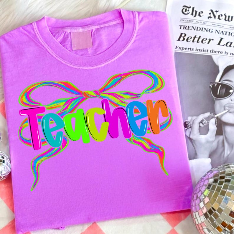 Op de afbeelding: Een lila t-shirt met het woord "Teacher" in kleurrijke bubbelletters. Een regenboogstrikontwerp staat boven het woord. Het shirt is opgevouwen en wordt op een oppervlak getoond.