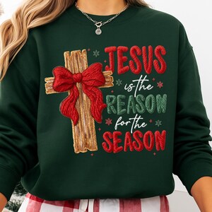 May include: Dark green sweatshirt with the text "JESUS is the REASON for the SEASON" in red and green. A wooden cross with a red bow is on the left side of the text. The sweatshirt is a festive holiday design.