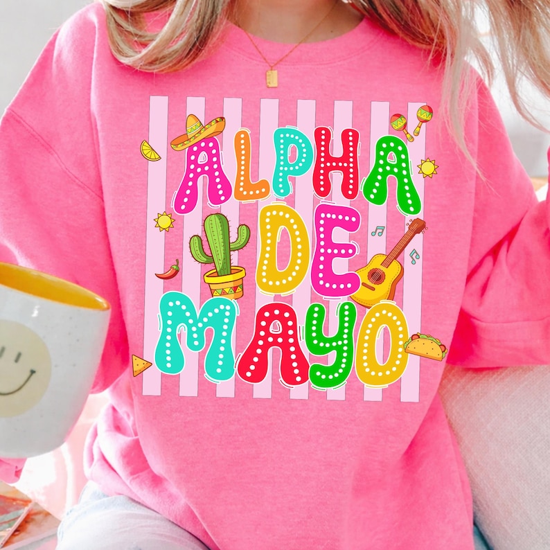 May include: A pink sweatshirt with the text "ALPHA DE MAYO" in colorful letters. The design includes a sombrero, cactus, guitar, maracas, and other festive graphics, perfect for a Cinco de Mayo celebration.