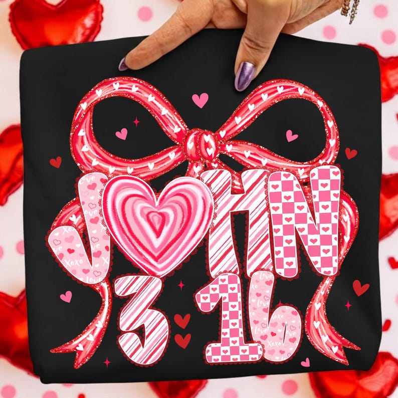 May include: Black t-shirt with a large red bow and the words "JOHN 3:16" in pink and white. The design includes heart shapes and a heart-shaped candy in the center. The shirt is held by a hand with a purple nail.