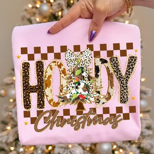 May include: A light pink sweatshirt with the word "HOODY" in a mix of leopard print and cowhide patterns. A Christmas tree with lights and a bow is in the center. The word "Christmas" is written below. Brown and gold accents.