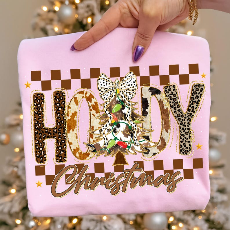 May include: A light pink sweatshirt with the word "HOODY" in a mix of leopard print and cowhide patterns. A Christmas tree with lights and a bow is in the center. The word "Christmas" is written below. Brown and gold accents.