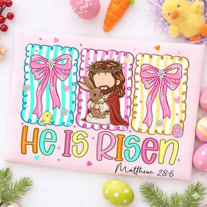 May include: A pink t-shirt with an Easter design, featuring the text "He Is Risen" and Matthew 28:6. The design includes Jesus with a bunny, bows, flowers, and an Easter egg. A small yellow chick is also featured.
