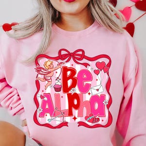 May include: Pale pink sweatshirt featuring a graphic with the words "Be Alpha" in red and pink. The design includes a cupid, hearts, a cupcake, and an envelope, framed by a red ribbon.