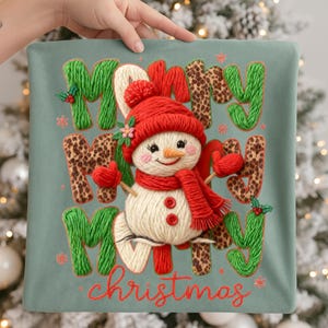 May include: A decorative Christmas pillow featuring a snowman design. The snowman wears a red hat, scarf, and mittens. The words "Merry Christmas" are embroidered in green, leopard print, and red on a teal background.