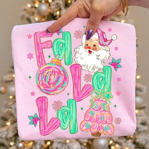 May include: A light pink sweatshirt with a festive design. The design includes the words "Fa La La La" in pink and green bubble letters, along with a Santa Claus illustration, a Christmas ornament, and a Christmas tree.
