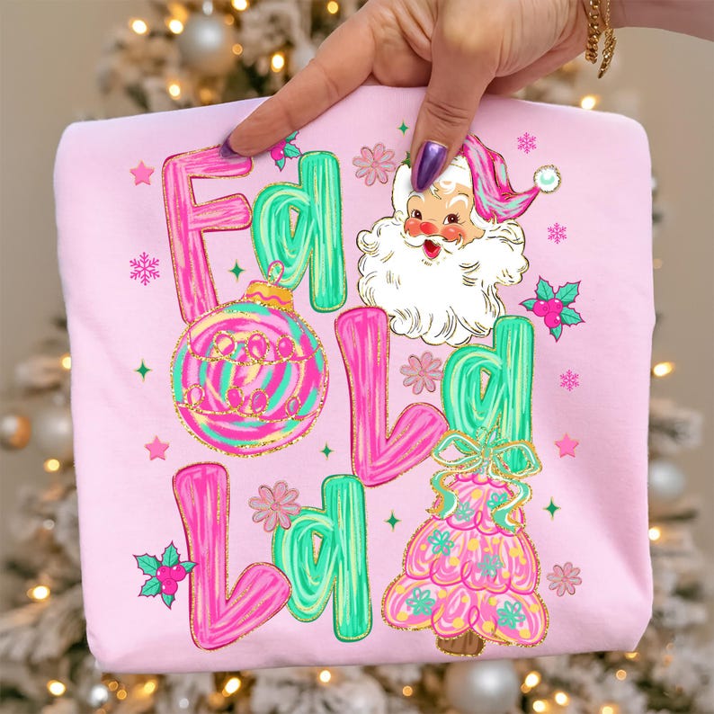 May include: A light pink sweatshirt with a festive design. The design includes the words "Fa La La La" in pink and green bubble letters, along with a Santa Claus illustration, a Christmas ornament, and a Christmas tree.