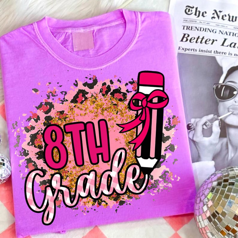 May include: A lilac-colored t-shirt with a graphic design for 8th grade. The design includes a pencil with a pink bow, leopard print accents, and the words "8th Grade" in pink and white. A newspaper is in the background.