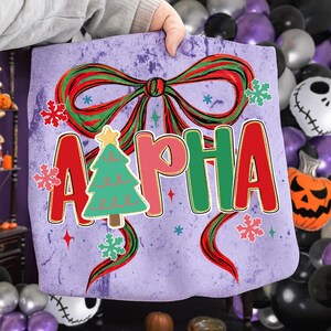 May include: A purple tote bag featuring a large red and green bow, snowflakes, and the word "ALPHA" in red, pink, and green letters. A green Christmas tree with a yellow star is in the center. The bag is held by a person.