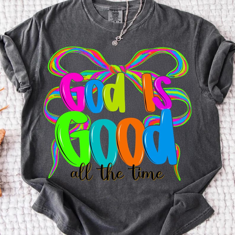 May include: Dark gray t-shirt with the words "God is Good" in bright, colorful letters. A rainbow-colored bow design is behind the text, and the phrase "all the time" is written below in gold.