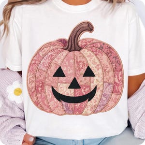 K&ouml;nnte beinhalten: Wei&szlig;es T-Shirt mit einem rosa Patchwork-K&uuml;rbis-Design. Der K&uuml;rbis hat ein l&auml;chelndes Jack-o'-Lantern-Gesicht und einen braunen Stiel. Das Design besteht aus verschiedenen Stoffen mit floralen Mustern. Ein Halloween-Themen-Grafik-T-Shirt.