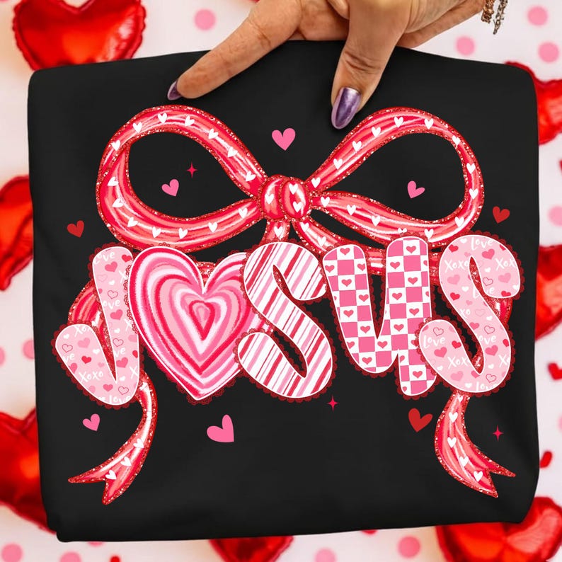 May include: Black t-shirt with a large, decorative bow and the word "JESUS" in pink and white. The letters are designed with hearts and patterns, with small hearts scattered around the design. A hand is holding the shirt.
