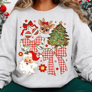 May include: A light grey sweatshirt with a festive Christmas design. The design includes a reindeer with a red bow, a Christmas tree, Santa Claus, red bows, and ornaments. The design is in red, green, and white.