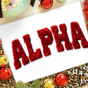 May include: A white surface with the word "ALPHA" spelled out in plush, red letters. The letters are thick and textured. The image is decorated with red and gold Christmas ornaments, a disco ball, and a leopard print fabric.