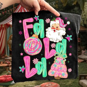 May include: Black t-shirt with a Christmas-themed design. The design features the words "F L O L L L O L" in pink and teal, along with a Santa Claus illustration, a Christmas tree, and ornaments.