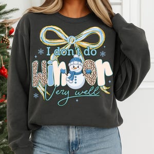 May include: Dark gray sweatshirt with a whimsical winter design. The text "I don't do winter very well" is displayed with a snowman graphic. The word "winter" has a leopard print pattern. A blue bow and snowflakes complete the design.