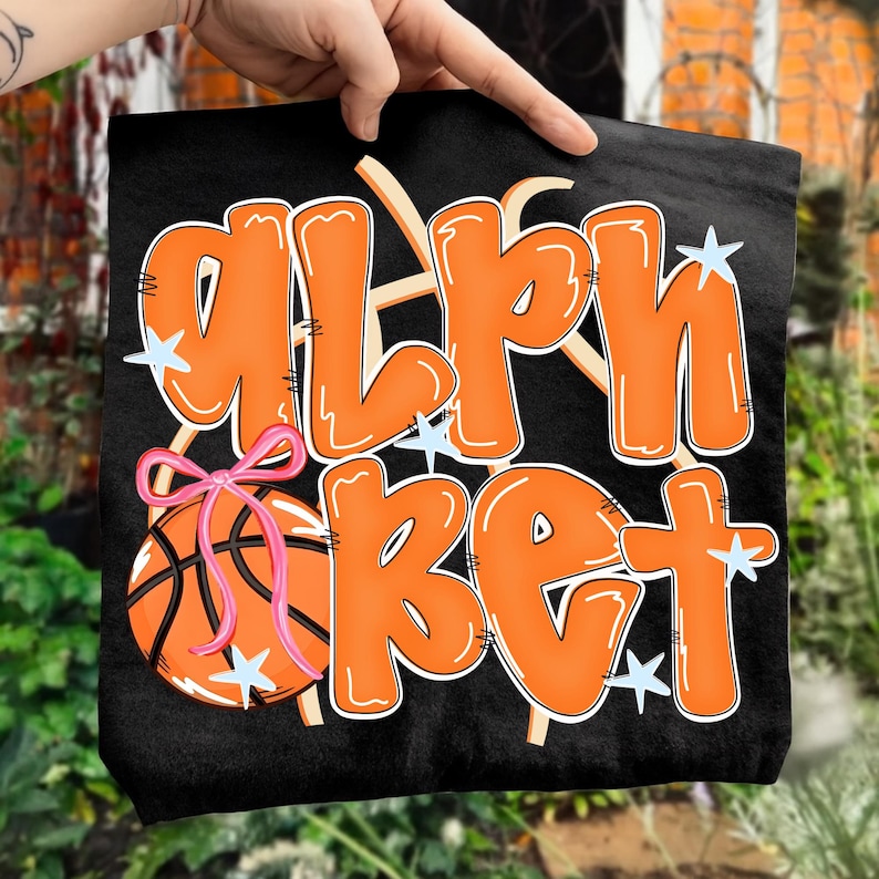 May include: Black tote bag featuring the word "ALPHABET" in orange bubble letters with white outlines. A basketball with a pink bow is on the left. Light blue star accents are scattered around the text.