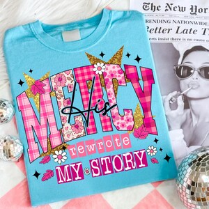 Op de afbeelding: Lichtblauw T-shirt met de tekst "MERCY His rewrote MY STORY" in roze, bloemen en gouden glitterletters. Het shirt heeft een ronde hals en korte mouwen. Bloemen en bladeren sieren het ontwerp.