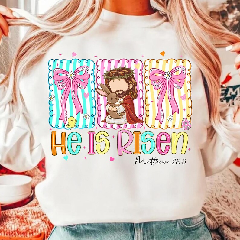 May include: White sweatshirt with a colourful graphic design. The design features three rectangular frames with a pink bow, a cartoon Jesus holding a bunny, and the text "He Is Risen" with "Matthew 28:6".
