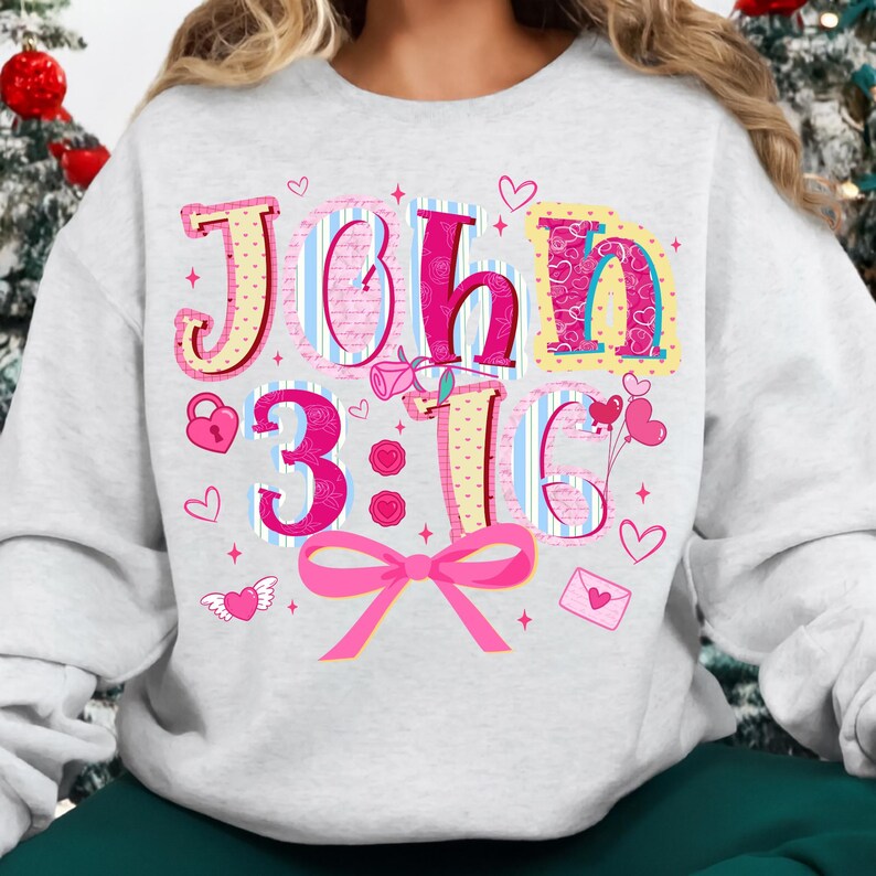 May include: A light grey sweatshirt featuring the text "John 3:16" in pink, yellow, and blue. The design includes hearts, a lock, a bow, and an envelope with a heart. The sweatshirt is a casual, comfortable style.