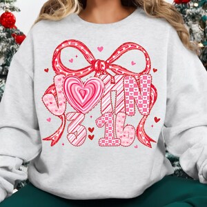 May include: A light gray sweatshirt with a large red bow and the text "JOHN 3:16" in pink and white. The design includes hearts and a heart-shaped candy in the center, perfect for Valentine's Day or a religious theme.