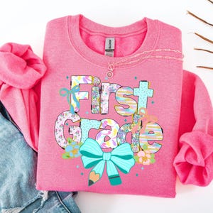 May include: Pink sweatshirt with the words "First Grade" in colorful, patterned letters and a large turquoise bow with a pencil graphic. The sweatshirt has long sleeves and a crew neck. A delicate necklace is draped across the top.