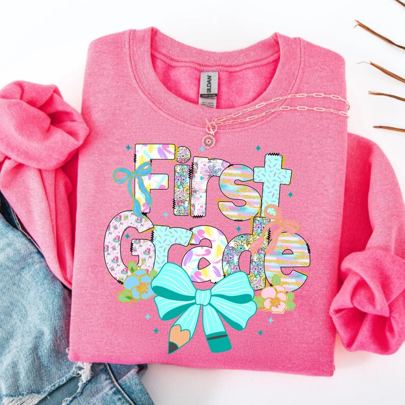 May include: Pink sweatshirt with the words "First Grade" in colorful, patterned letters and a large turquoise bow with a pencil graphic. The sweatshirt has long sleeves and a crew neck. A delicate necklace is draped across the top.