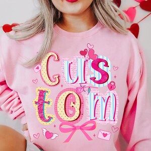 May include: Pink sweatshirt with the word "CUSTOM" in a colorful, decorative font. The letters are adorned with hearts, bows, and other Valentine's Day themed graphics. The sweatshirt is a light pink color.