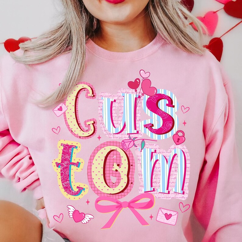 May include: Pink sweatshirt with the word "CUSTOM" in a colorful, decorative font. The letters are adorned with hearts, bows, and other Valentine's Day themed graphics. The sweatshirt is a light pink color.