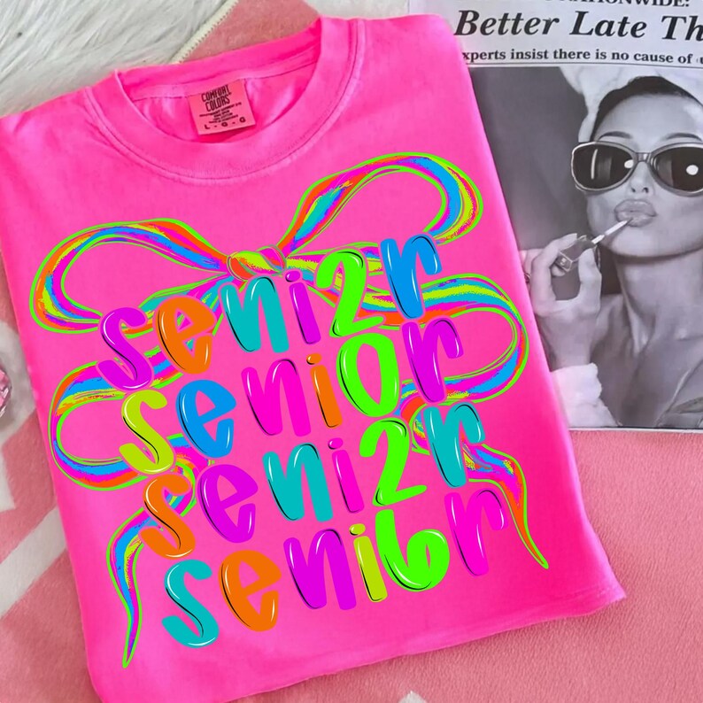May include: Bright pink t-shirt with the word "Senior" in colorful, layered text. A large, rainbow-colored bow design arches over the text. The shirt has a crew neck and short sleeves. The Comfort Colors brand tag is visible.