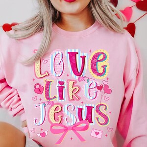 May include: Pink sweatshirt with the words "LOVE LIKE JESUS" in a colorful, whimsical font. The design includes hearts, a lock, a bow, and other decorative elements, perfect for Valentine's Day or expressing faith.