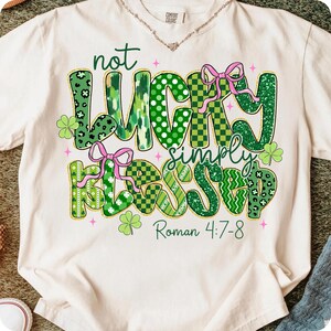 May include: Cream-colored t-shirt with a St. Patrick's Day design. The graphic reads "not LUCKY simply BLESSED" in green, with pink bows and shamrocks. The text "Roman 4:7-8" is below the main text.