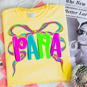 May include: Yellow t-shirt with a colorful bow design and the word "PAPA" in bright pink, teal, and green. The shirt is folded and displayed on a pink and white striped surface.