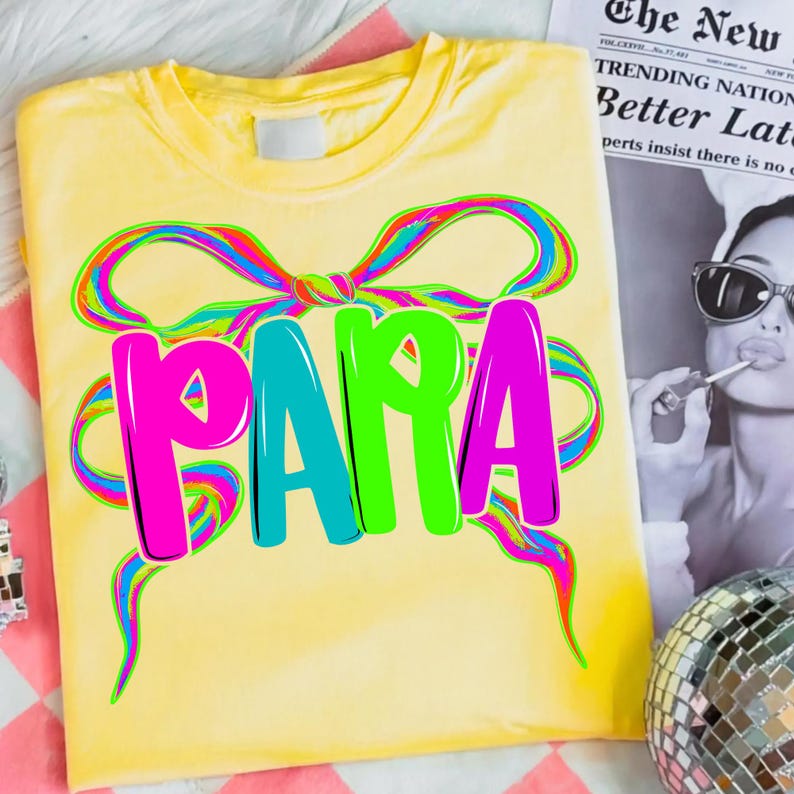 May include: Yellow t-shirt with a colorful bow design and the word "PAPA" in bright pink, teal, and green. The shirt is folded and displayed on a pink and white striped surface.